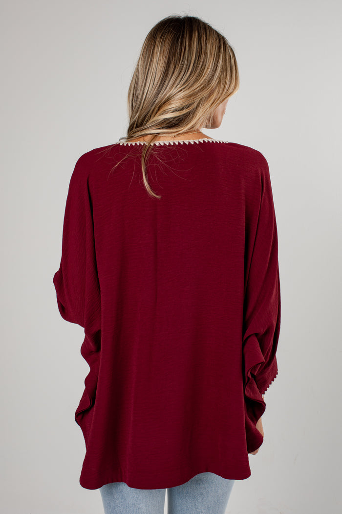A woman viewed from behind wearing a loose, deep burgundy long-sleeve top with decorative crochet trim around the neckline and sleeve hems, paired with light blue jeans against a plain light gray background.
