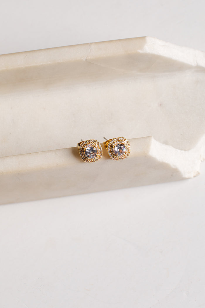 A pair of gold square stud earrings with clear stones, resting on a light beige stone block against a white background.