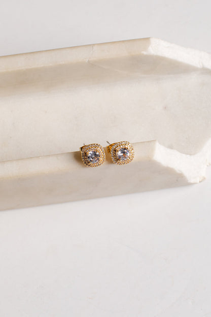 A pair of gold square stud earrings with clear stones, resting on a light beige stone block against a white background.