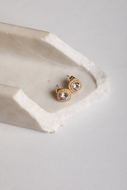 A pair of gold stud earrings with clear round gemstones, displayed on a textured white stone tray against a light gray background.