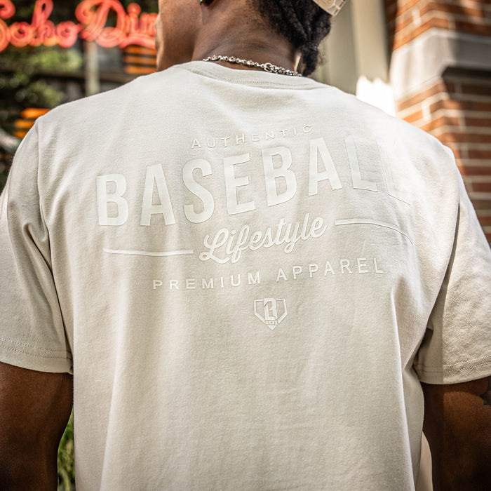 Person wearing a beige t-shirt with 'Authentic Baseball Lifestyle Premium Apparel' text on the back, standing outdoors