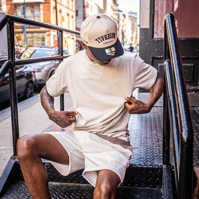 Person wearing a beige 'Authentic Baseball Lifestyle PREMIUM APPAREL' t-shirt and shorts with a cap, sitting on a metal staircase.