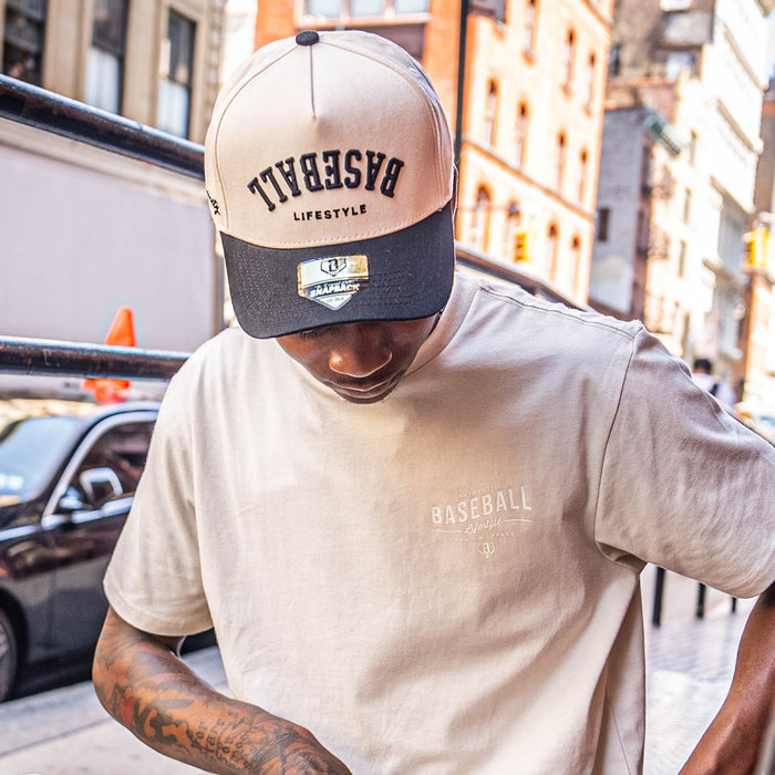 Man wearing a beige and navy cap and matching t-shirt with 'Authentic Baseball Lifestyle PREMIUM APPAREL' text, standing on a city street.