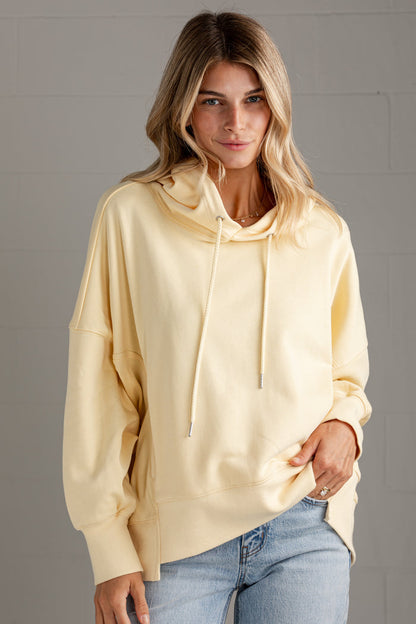 A woman wearing a pale yellow oversized pullover hoodie with drawstrings and front pouch pockets, paired with light blue jeans, standing against a gray paneled wall.