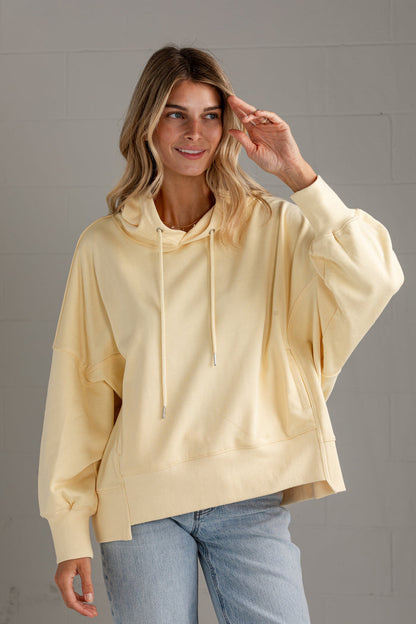 A woman wearing a pale cream-colored oversized pullover hoodie with balloon sleeves and drawstrings, paired with light blue jeans, standing against a gray brick wall.