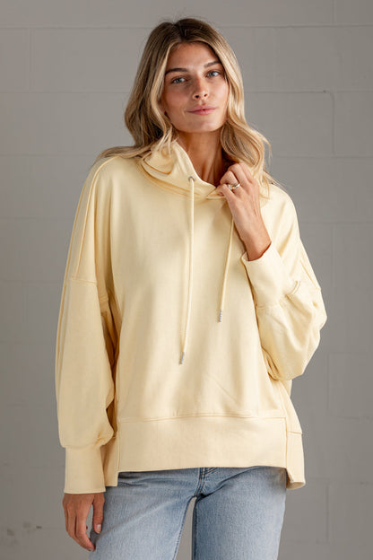 A woman wearing a pale yellow oversized pullover hoodie with drawstrings and long sleeves, paired with light blue jeans, standing against a gray brick wall.