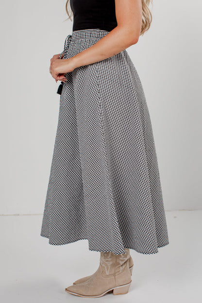 A woman wearing a high-waisted, A-line midi skirt with a black and white micro-check pattern, styled with a black top and beige cowboy boots against a plain white studio background.
