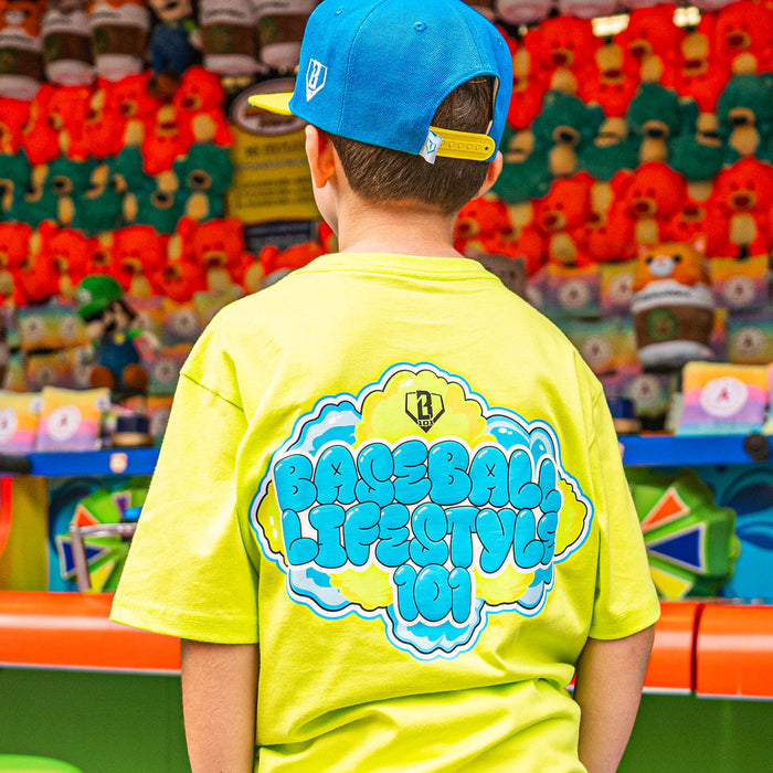 Child wearing a bright yellow t-shirt with blue and green 'Baseball Lifestyle' text, standing in front of toy store shelves.