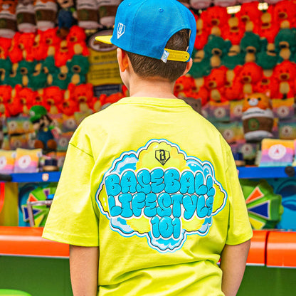 Child wearing a bright yellow t-shirt with blue and green 'Baseball Lifestyle' text, standing in front of toy store shelves.