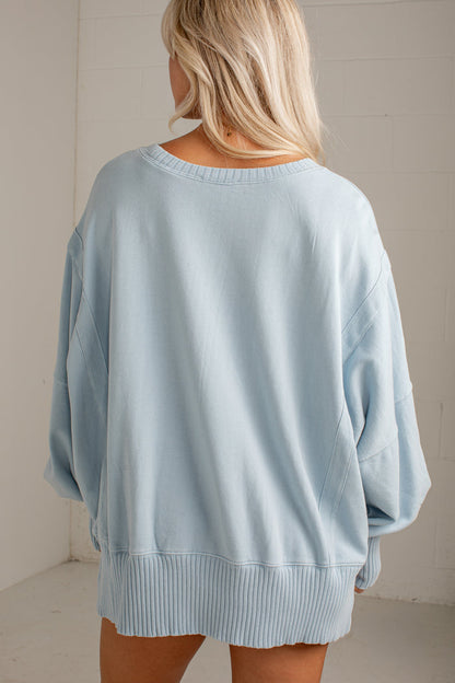 A woman viewed from behind wearing an oversized light blue knit sweater with wide sleeves and a ribbed hem, set against a plain off-white wall.
