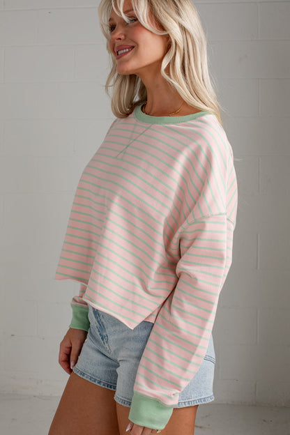 A woman wearing a pastel pink and mint green striped long-sleeve crop sweatshirt with solid green ribbed cuffs and collar, paired with denim shorts, standing against a white brick wall.