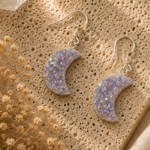 Crescent moon-shaped earrings with glittery finish on a textured beige surface with dried flowers.