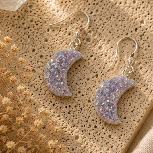 Crescent moon-shaped earrings with glittery finish on a textured beige surface with dried flowers.