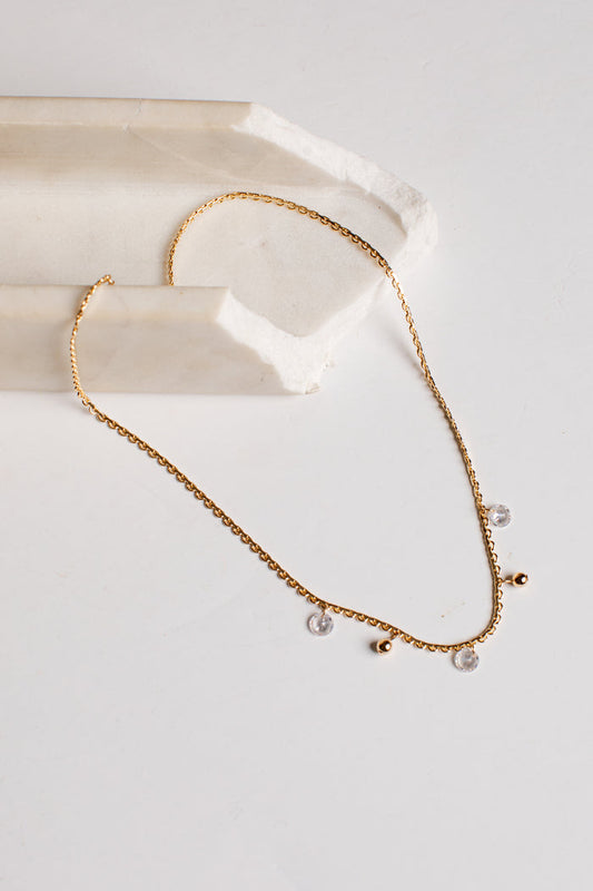 A delicate gold chain necklace with small clear crystal pendants and tiny gold beads, displayed on a white surface next to a cream-colored stone prop.