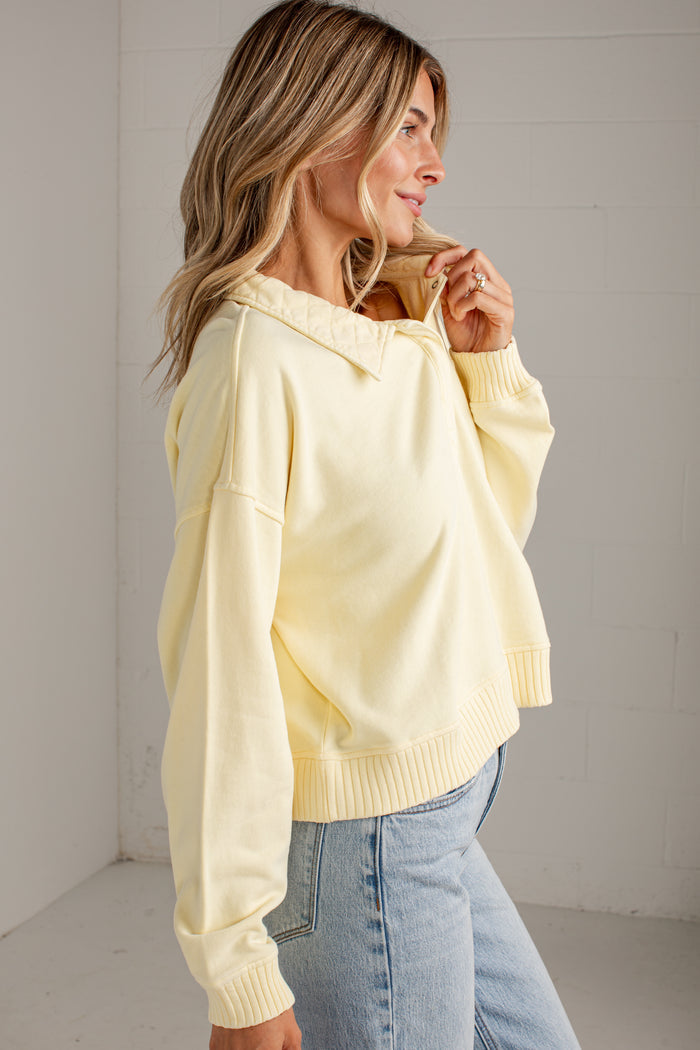 A woman wearing a pale yellow oversized knit sweater with a wide collar and ribbed cuffs, paired with light blue jeans, standing in a bright white room.