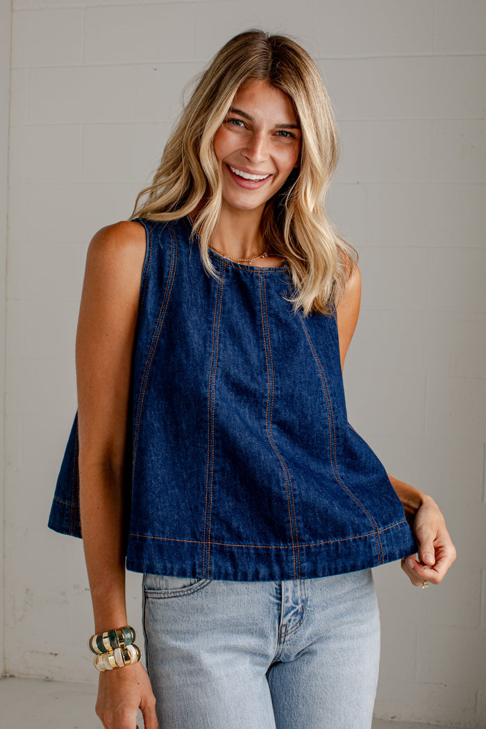 A woman wearing a sleeveless dark blue denim top with light-wash jeans, standing against a light gray wall.