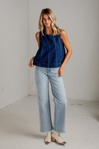 A woman modeling a sleeveless dark blue denim top paired with light-wash wide-leg jeans and beige heeled sandals, standing in a bright white studio space.