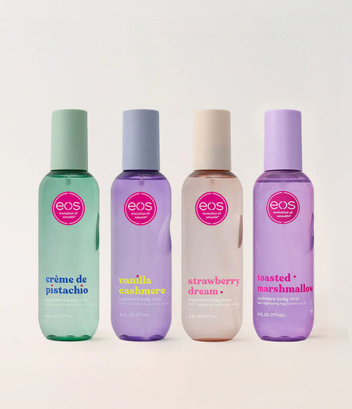 Four EOS Cosmetics body care bottles lined up on a light beige background: crème de pistachio in mint green, vanilla soapsake in lavender, strawberry dream in pale peach, and toasted marshmallow in light purple. Each bottle has a pink circular EOS logo and the scent name printed below.