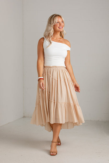 A woman modeling a white one-shoulder top and a tiered beige midi skirt, standing in a bright, minimalist studio with white brick walls.