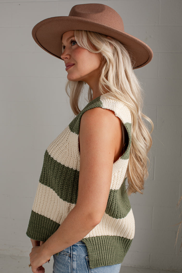 A woman wearing a brown wide-brim hat and a sleeveless knit vest with horizontal olive green and cream stripes, paired with blue jeans, photographed against a light gray brick wall.