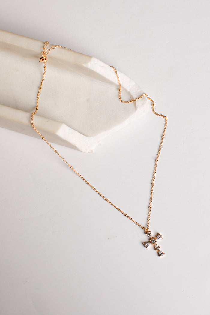 A delicate rose gold chain necklace with a small cross pendant, draped over a white stone display block on a light gray background.