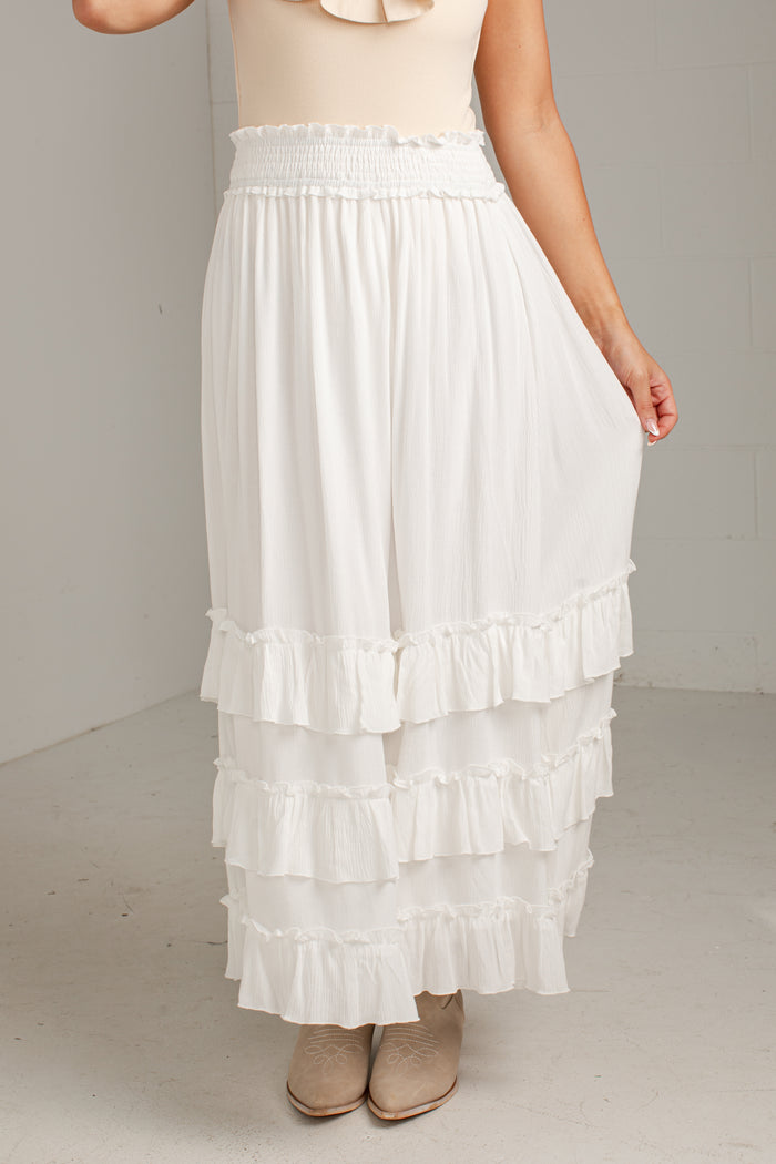 A white tiered maxi skirt with a smocked elastic waistband, shown on a model wearing beige ankle boots against a neutral studio background.