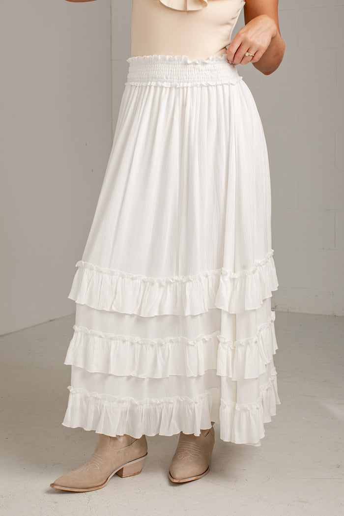 A white tiered maxi skirt with an elastic smocked waistband, shown on a model wearing beige ankle boots against a neutral studio background.