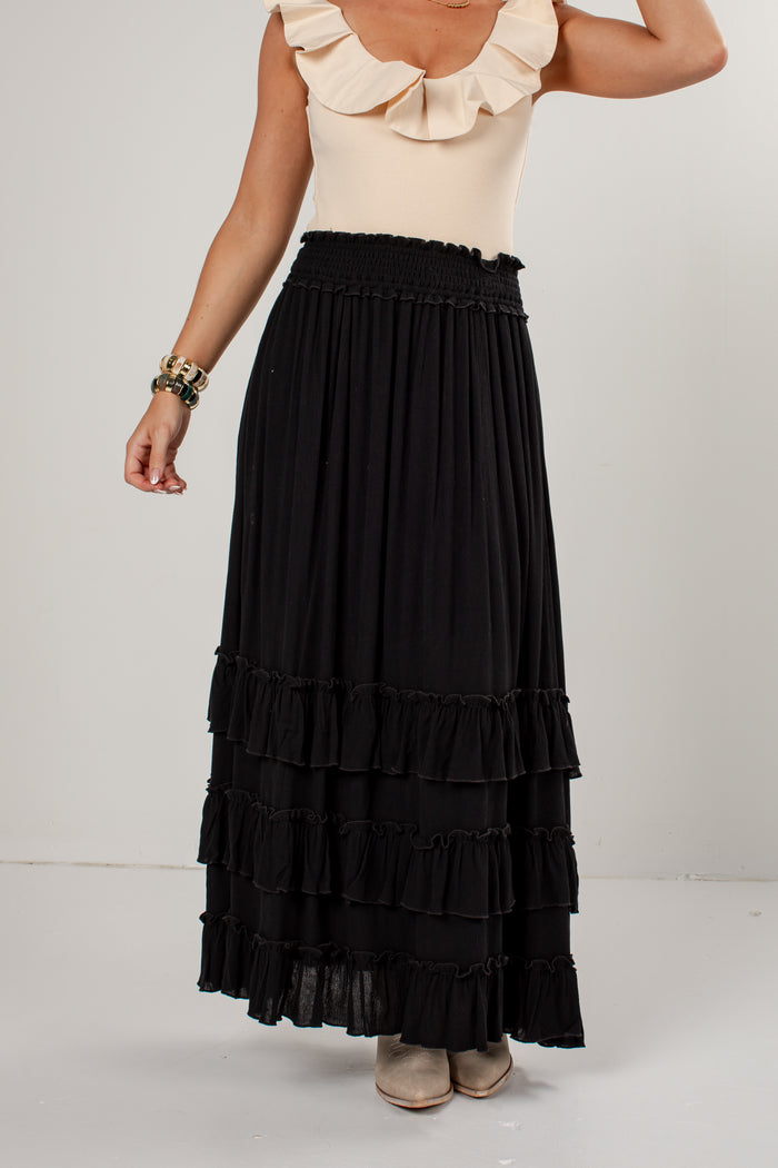 A model wearing a long black tiered maxi skirt with a smocked waistband, paired with a cream ruffled top and beige shoes, shown from the chest down against a plain light gray studio background.