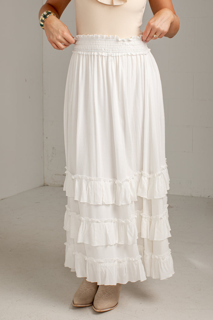 A white tiered maxi skirt with a smocked elastic waistband, shown on a model wearing beige boots and a cream top in a neutral studio setting.