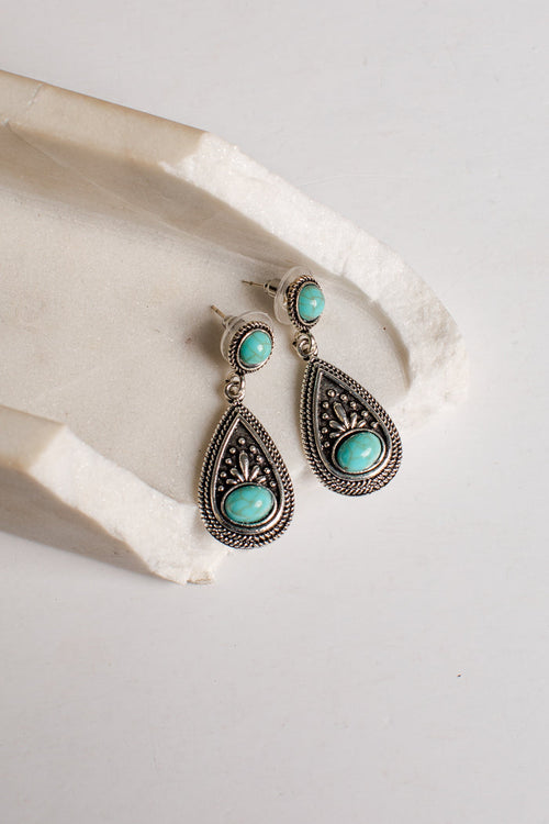 A pair of silver-toned teardrop dangle earrings with oval turquoise stones, displayed on a textured white stone prop against a light neutral background.