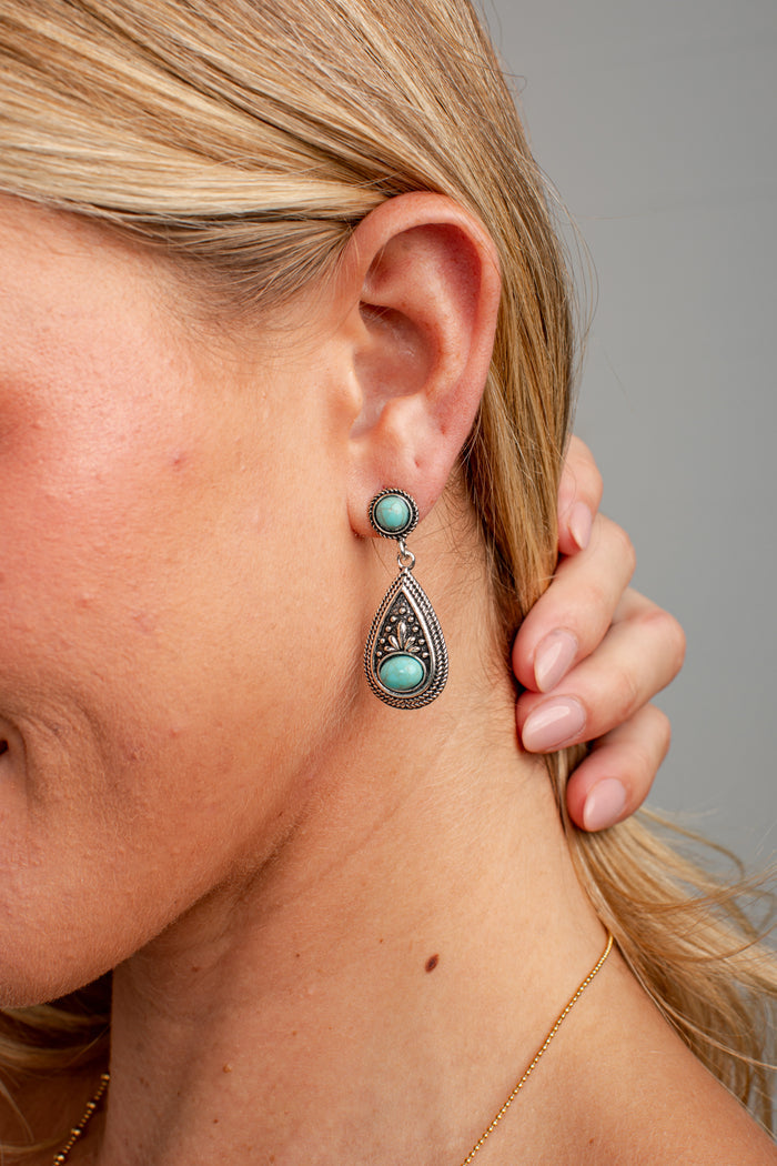 A close-up of a woman's ear wearing a silver-toned drop earring with a round turquoise stone and a teardrop-shaped silver filigree pendant with a turquoise center.