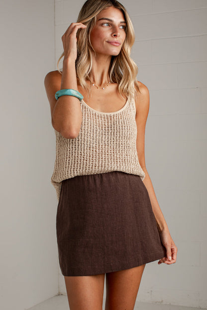 A woman modeling a beige chunky knit tank top and a brown linen mini skirt against a white paneled wall.