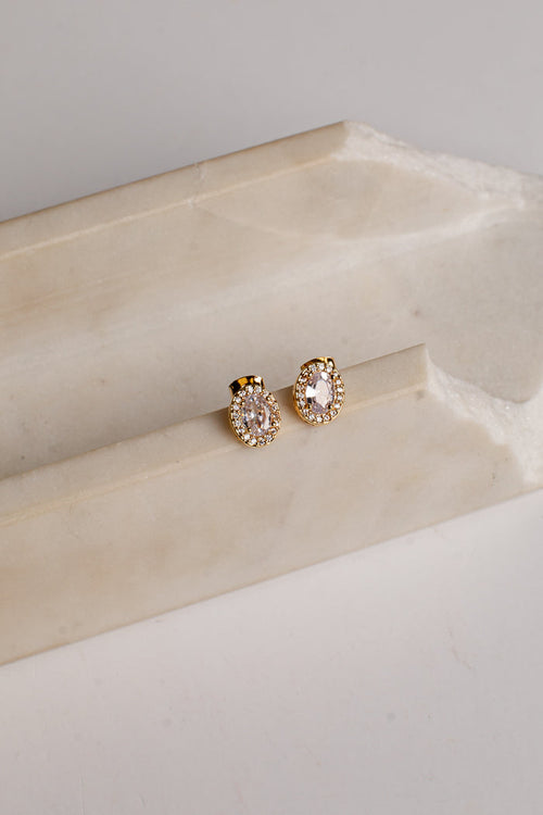 A pair of gold-toned stud earrings with oval clear gemstones and small surrounding clear stones, displayed on a cream-colored stone riser against a light neutral background.