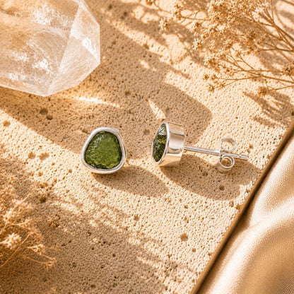 A pair of silver stud earrings with Moldavite, displayed on a textured beige surface with dried flowers and a clear crystal.
