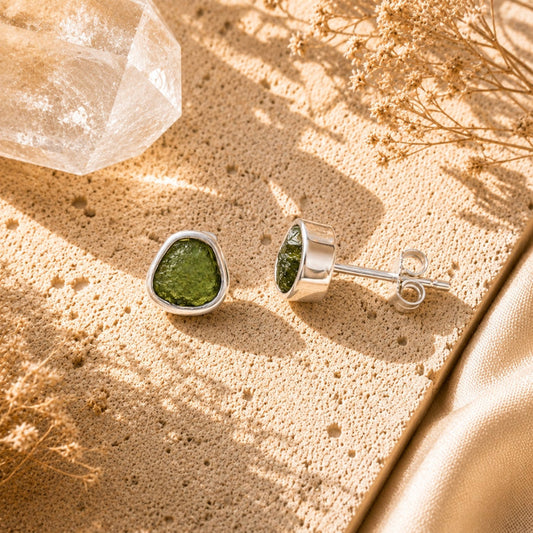 A pair of silver stud earrings with Moldavite, displayed on a textured beige surface with dried flowers and a clear crystal.