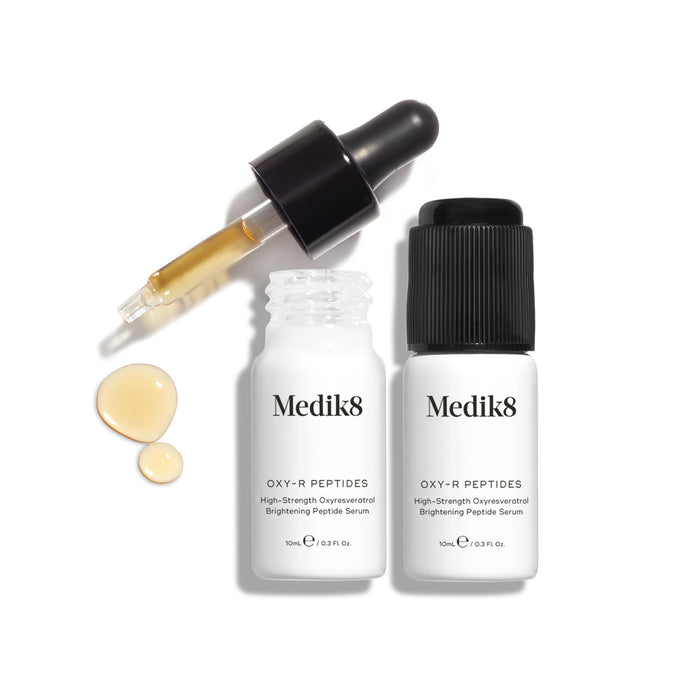 Two small white bottles of Oxy-R Peptides serum are displayed. A black dropper tool lies nearby, and two drops of yellow serum are visible on the white surface