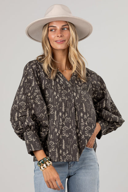 A woman wearing a dark brown and beige floral print blouse with voluminous balloon sleeves, paired with light blue jeans and a beige wide-brim hat, against a solid light gray background.