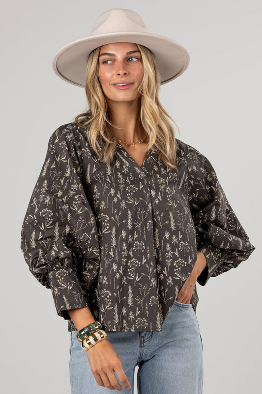 A woman wearing a dark brown and beige floral print blouse with voluminous balloon sleeves, paired with light blue jeans and a beige wide-brim hat, against a solid light gray background.