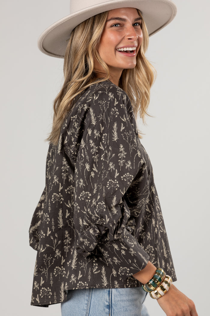 A woman wearing a dark brown long-sleeve blouse with a beige botanical floral pattern, styled with a wide-brimmed cream hat and light blue jeans against a light gray studio background.