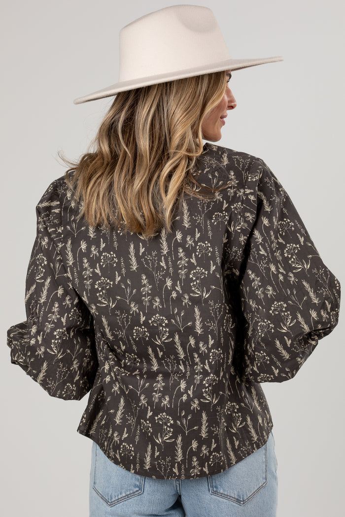 A woman shown from the back wearing a dark brown long-sleeve blouse with a beige botanical pattern, paired with light blue jeans and a cream wide-brim hat, against a light gray studio background.