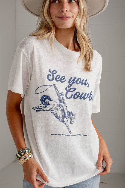 A woman wearing a white graphic t-shirt with a blue cowboy design and the text 'See you, Cowboy!' paired with light-wash jeans and a wide-brimmed white hat, standing against a light gray brick wall.