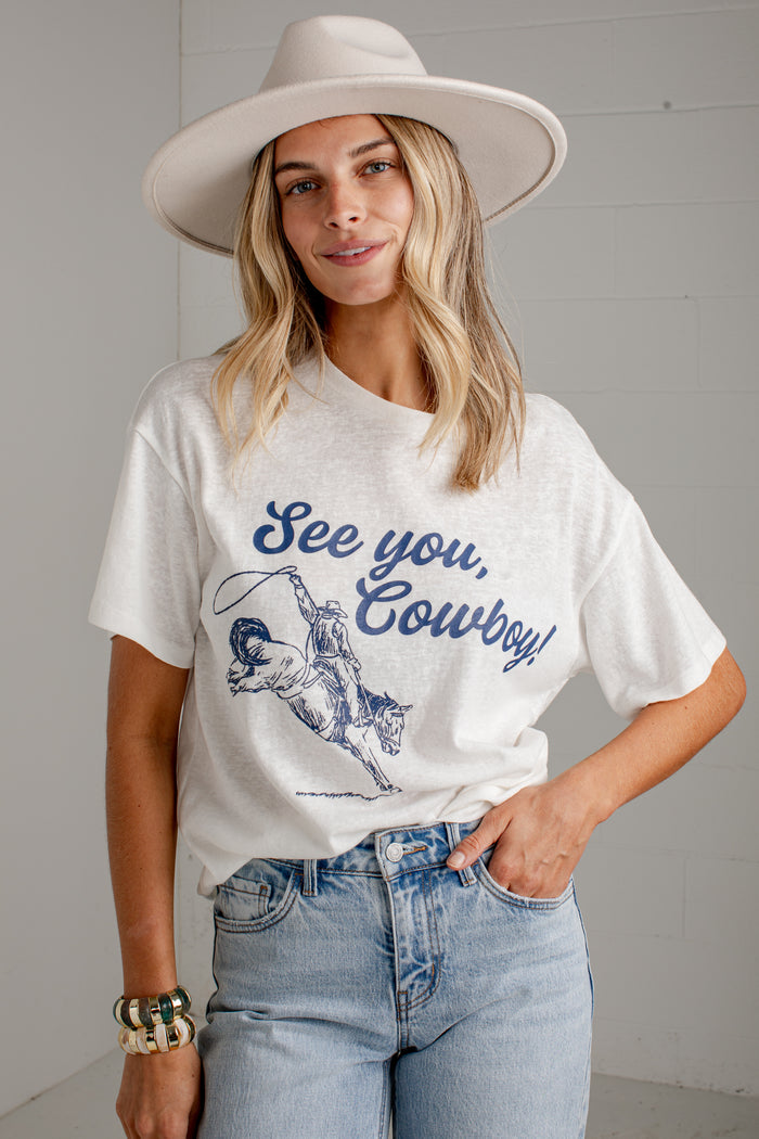 A woman wearing a white graphic t-shirt with a blue cowboy design and the text 'See you, Cowboy!' paired with light-wash jeans and a wide-brimmed white hat, standing against a light gray brick wall.