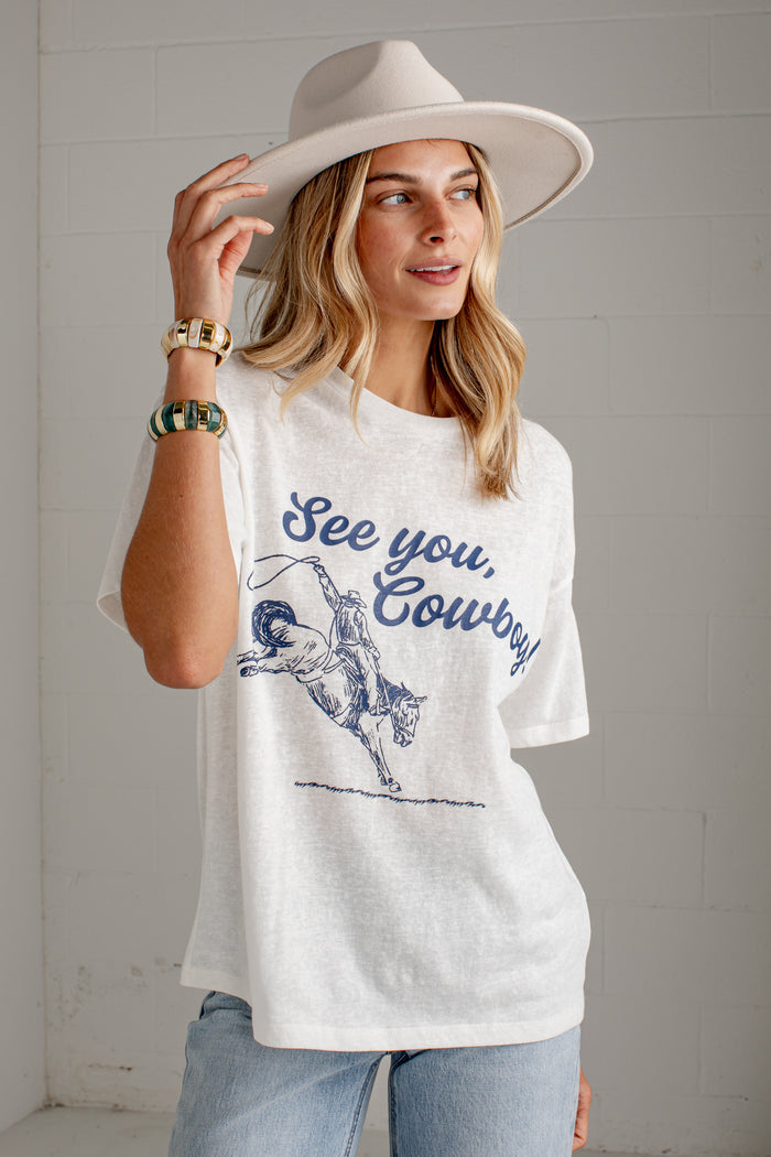 A woman wearing a white graphic t-shirt with a blue 'See you, Cowboy' print and a cowboy illustration, paired with a wide-brimmed beige hat and light blue jeans, standing against a white brick wall.