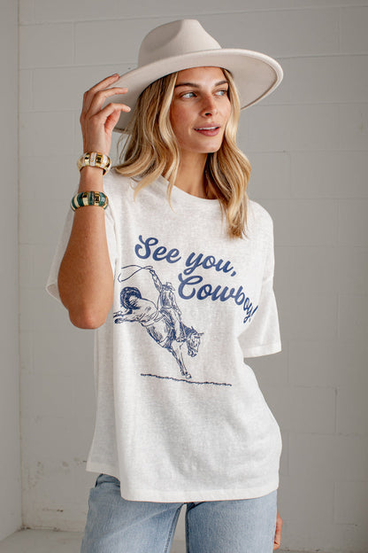 A woman wearing a white graphic t-shirt with a blue 'See you, Cowboy' print and a cowboy illustration, paired with a wide-brimmed beige hat and light blue jeans, standing against a white brick wall.