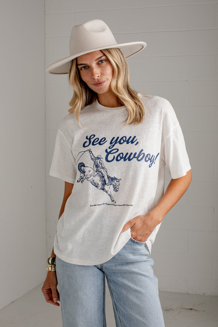 A woman wearing a white graphic t-shirt with a blue 'See you, Cowboy!' print, paired with light blue jeans and a wide-brimmed beige hat, standing against a white paneled wall.