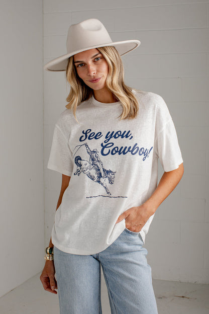 A woman wearing a white graphic t-shirt with a blue 'See you, Cowboy!' print, paired with light blue jeans and a wide-brimmed beige hat, standing against a white paneled wall.