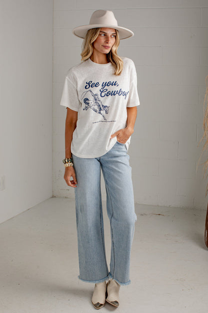 A woman wearing a white graphic t-shirt with a blue cowboy design and the text 'See you, Cowboy!' paired with light-wash jeans and a wide-brimmed white hat, standing against a light gray brick wall.