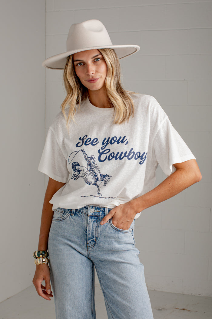 A woman wearing a white graphic t-shirt with a blue 'See you, Cowboy' print, paired with light-wash jeans and a wide-brimmed cream hat, standing against a white paneled wall.