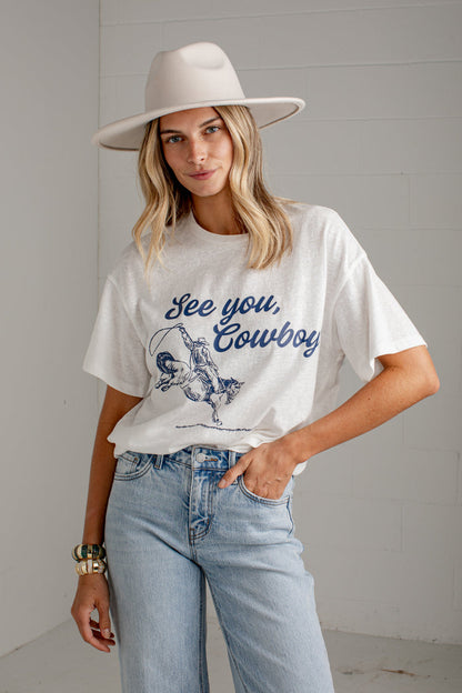 A woman wearing a white graphic t-shirt with a blue 'See you, Cowboy' print, paired with light-wash jeans and a wide-brimmed cream hat, standing against a white paneled wall.