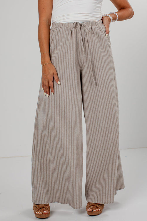 A person wearing wide-leg pleated pants in a taupe-beige color with thin vertical stripes, paired with a white top and brown sandals, shown from the waist down against a light gray studio background.